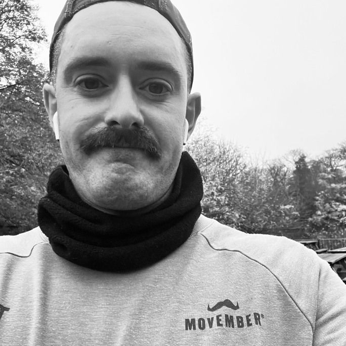Black and white photo of handsome man, sporting an impressive moustache, exercising outdoors and smiling to camera.