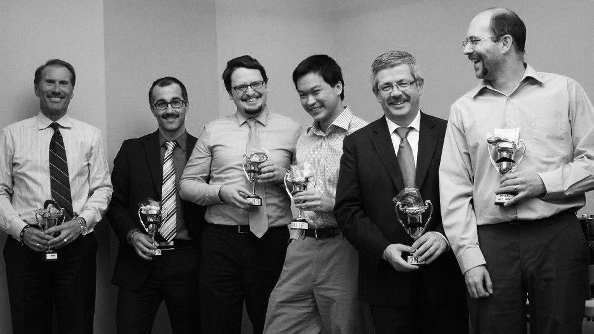 A group of colleagues in suit and tie, all sporting moustaches, hold trophies that also have moustaches on them